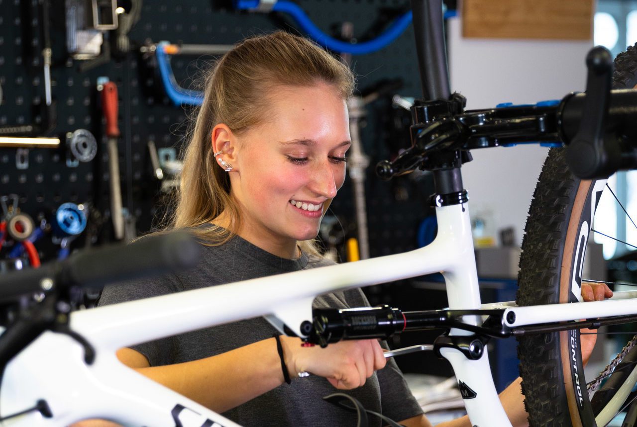 Jana Blickenstorfer, eine junge Frau, Anfang Zwanzig, blond und strahlen lächelnd steht in der Fahrrad Werkstatt hinter einem weissen Mountainbike von Specialized.